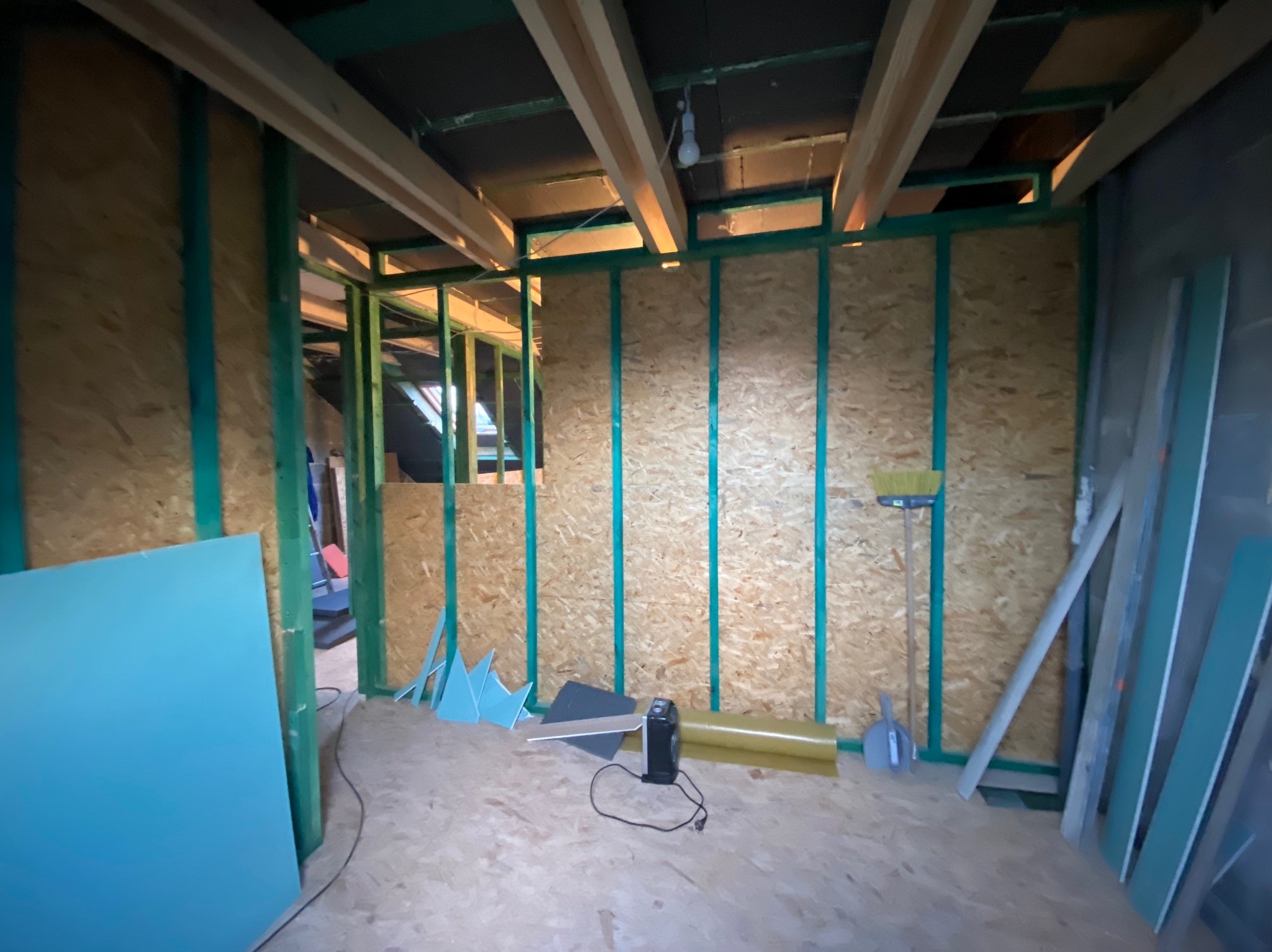 Wnętrze w trakcie adaptacji poddasza: widoczne drewniane belki stropowe, ściany obłożone płytą OSB z zielonymi listwami, płyty kartonowo-gipsowe i narzędzia pracy.