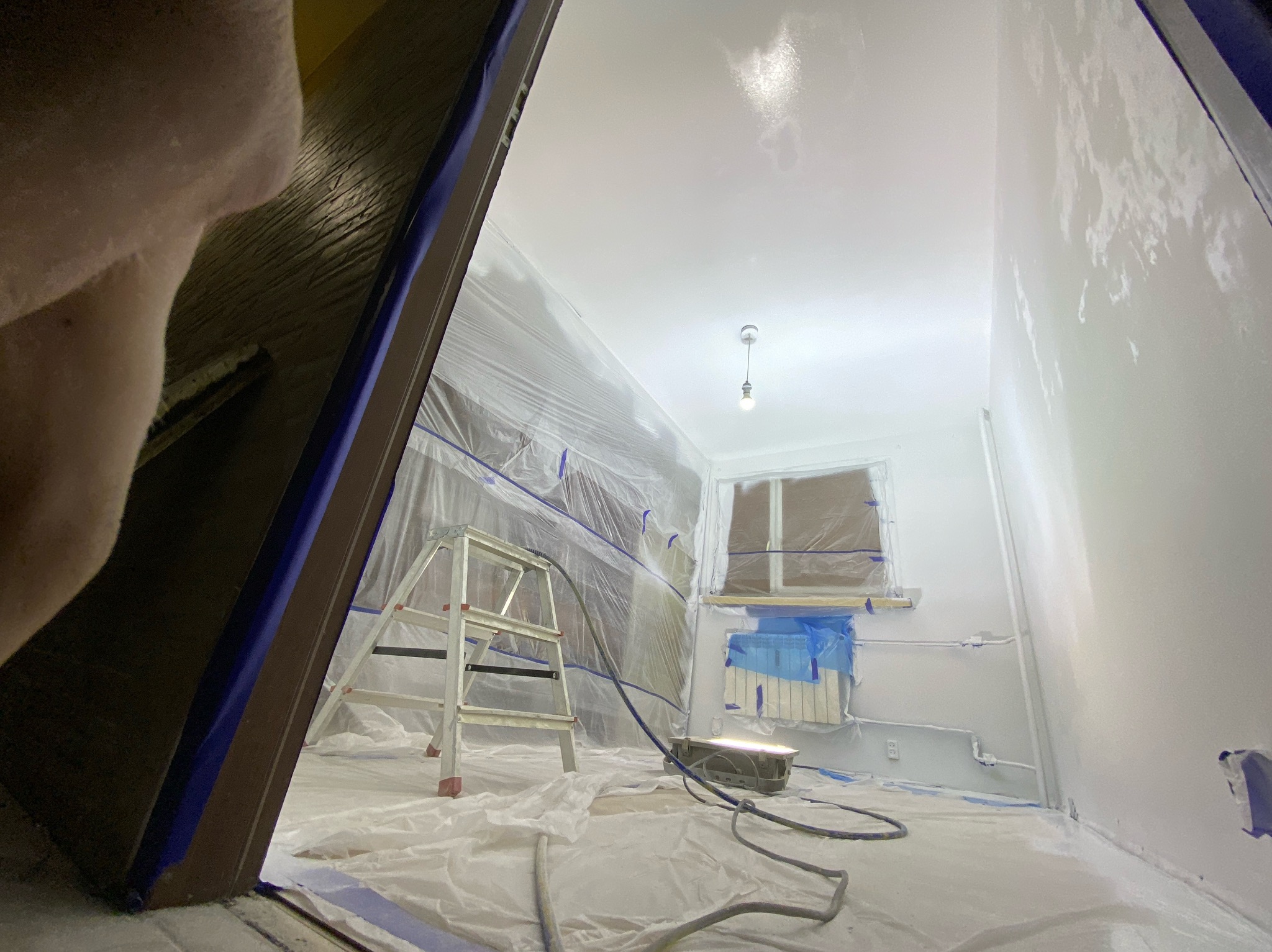 Pokój przygotowany do malowania: folia ochronna na podłodze i meblach, drabina, widoczne ślady świeżo nałożonej farby na ścianach.