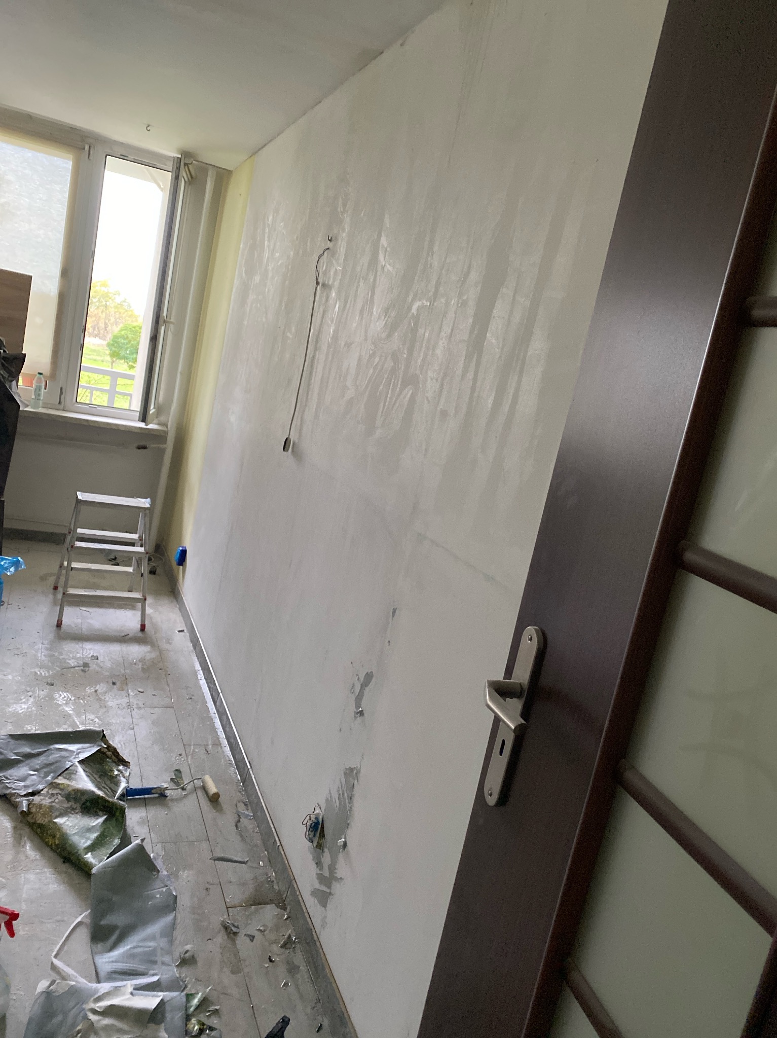 Ściana w trakcie szpachlowania z widocznymi śladami po taśmie malarskiej, podłoga zabezpieczona folią, drabina i fragment drzwi z klamką w tle.