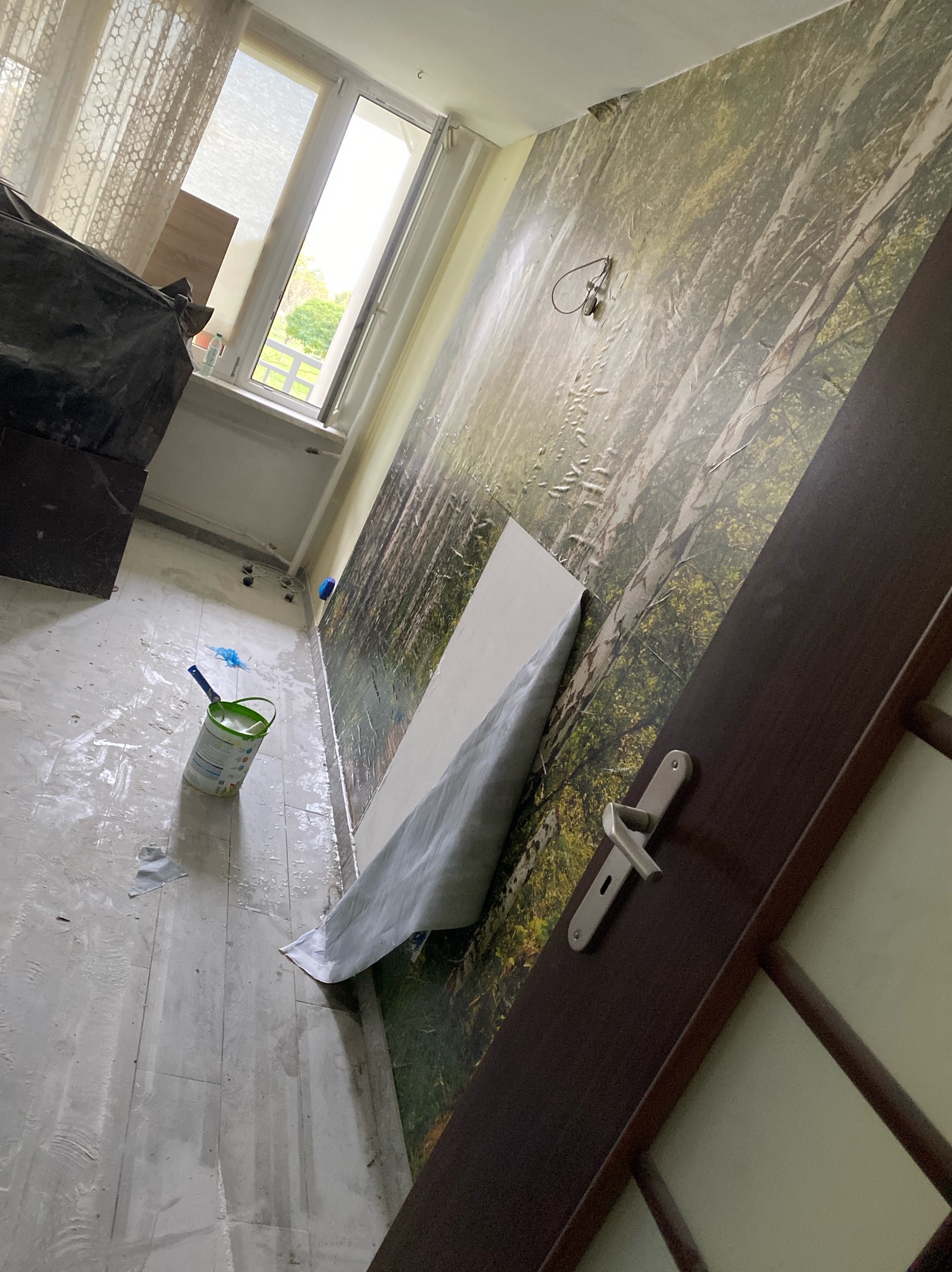 Pokój w trakcie odświeżania: ściana z fototapetą leśną częściowo zabezpieczona folią malarską, puszka farby i pędzel na podłodze, widoczny fragment ciemnych drzwi.
