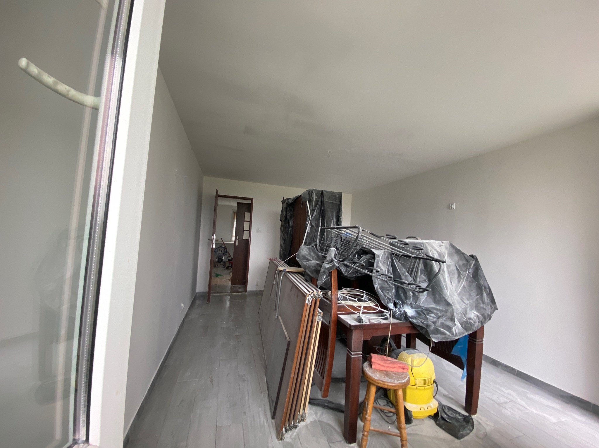 Pokój po świeżym malowaniu, z widocznymi śladami prac: zabezpieczone meble pod folią, drabina, stolik i krzesło robocze, oraz fragment odnowionego okna.