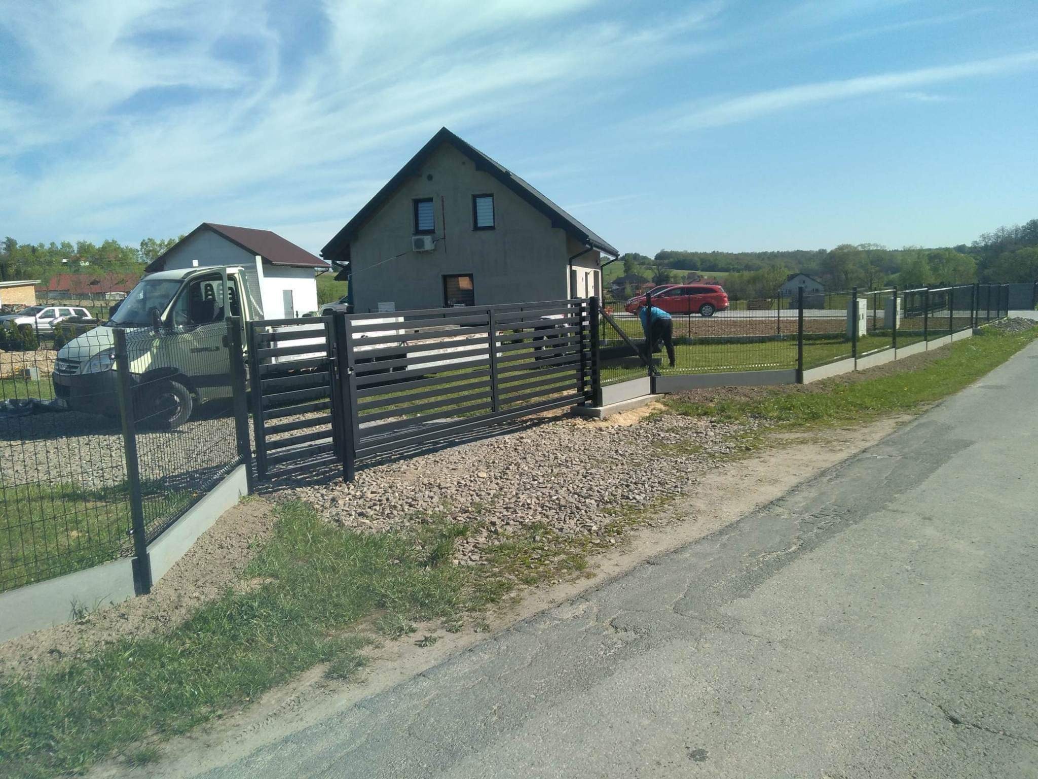 Montaż czarnego ogrodzenia panelowego z podmurówką betonową, widoczny pracownik w niebieskiej koszulce, samochód dostawczy i dom jednorodzinny w tle.