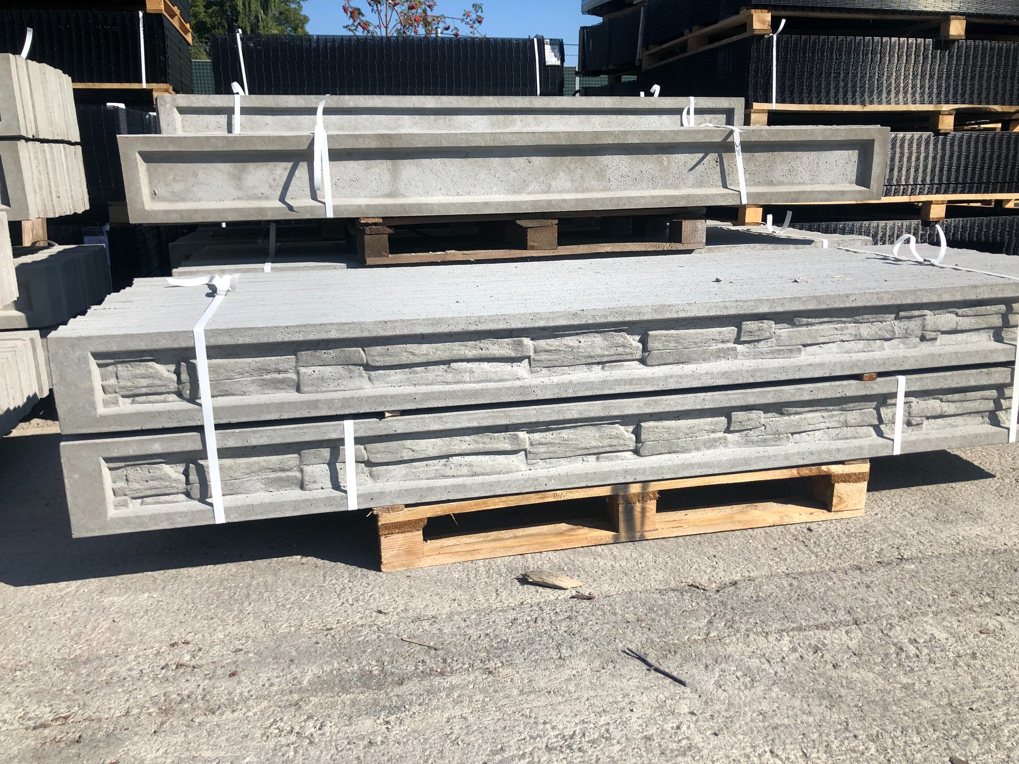 Stos betonowych elementów ogrodzeniowych na paletach, z widoczną fakturą imitującą kamień na niektórych płytach, zabezpieczonych białymi taśmami.