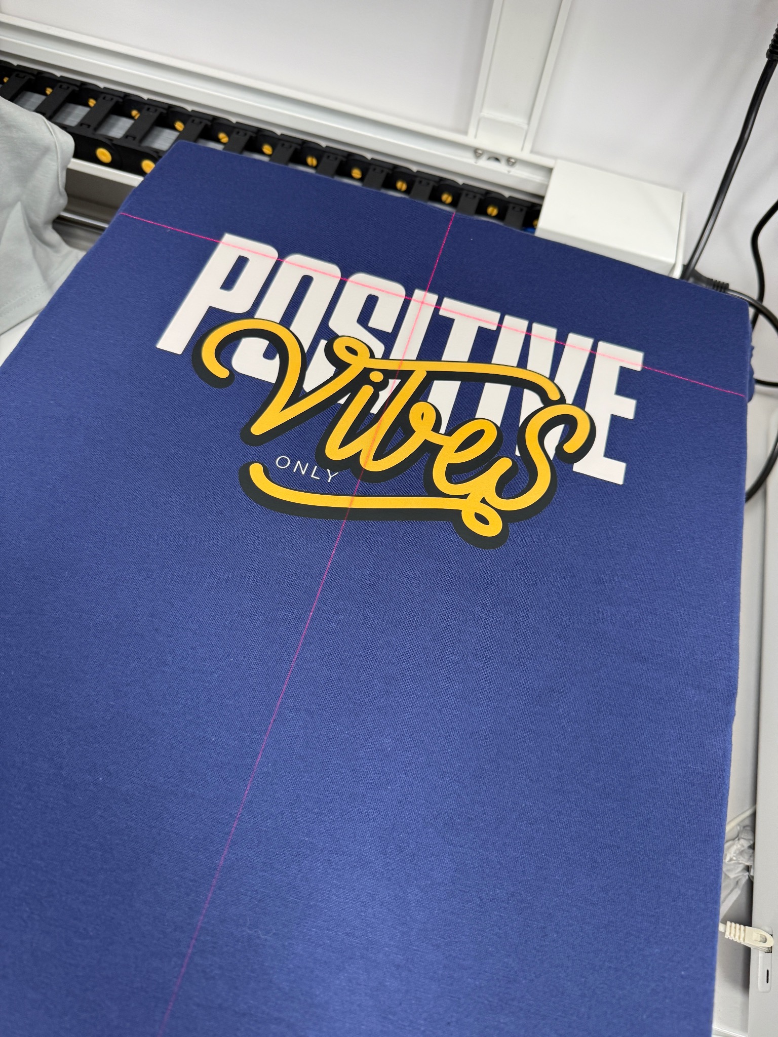 Niebieska koszulka z nadrukiem 'Positive Vibes Only', umieszczona w maszynie do nadruków, z widocznymi laserowymi liniami pozycjonującymi.