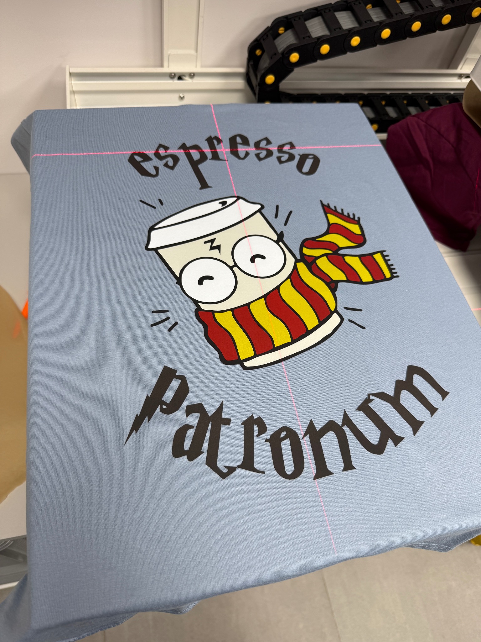 Jasnoniebieska koszulka z nadrukiem: kubek kawy w szaliku, napis 'espresso patronum'. Widoczne linie lasera drukarki DTG.