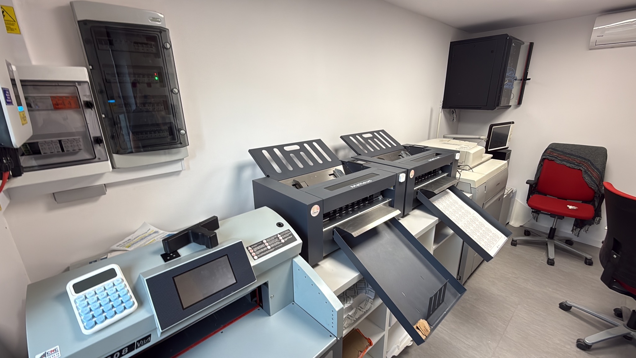Wnętrze drukarni z maszynami do cięcia i druku. Widoczne panele elektryczne na ścianie, biurko z kalkulatorem i krzesło biurowe. Ujęcie z perspektywy szerokokątnej.
