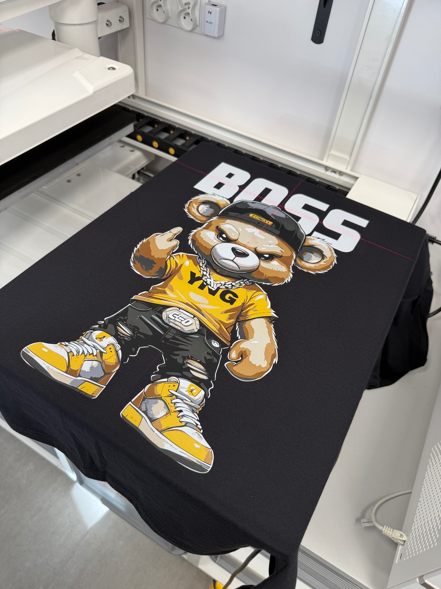 Czarna koszulka z nadrukiem kreskówkowego misia 'BOSS' w czapce i żółtej koszulce, leżąca na stole drukarki DTG z widocznymi laserowymi znacznikami.