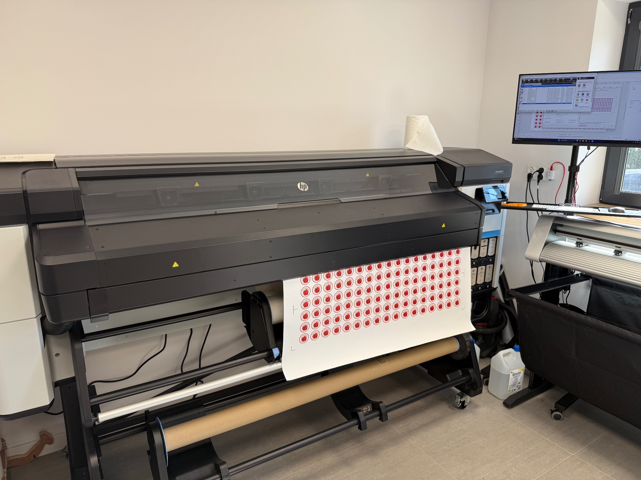 Drukarka wielkoformatowa HP w Jatne drukuje arkusz naklejek z czerwonymi okręgami. Widoczny monitor z oprogramowaniem drukarki i rolka papieru. Profesjonalny wydruk.