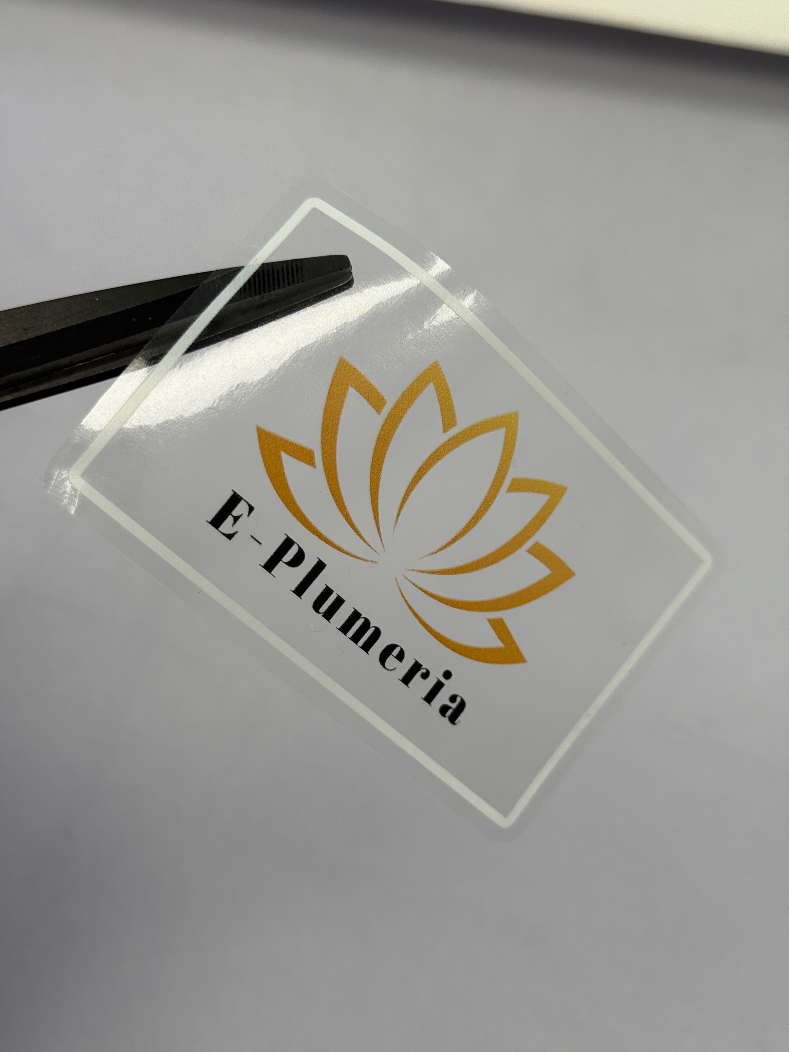 Przezroczysta naklejka z logo 'E-Plumeria' w kolorze złotym i czarnym, częściowo przytrzymywana przez czarne pęsety, ukazująca precyzyjne cięcie po obrysie.
