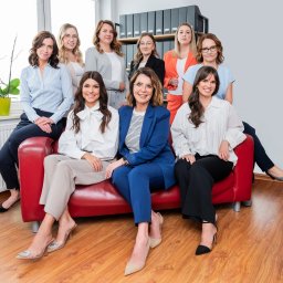 SUKURS SPÓŁKA Z OGRANICZONĄ ODPOWIEDZIALNOŚCIĄ - Grupowe portretowe zdjęcie dziewięciu uśmiechniętych kobiet w eleganckich strojach, siedzących na czerwonej sofie w biurze z regałem pełnym segregatorów w tle.