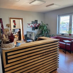 SUKURS SPÓŁKA Z OGRANICZONĄ ODPOWIEDZIALNOŚCIĄ - Nowoczesna recepcja z drewnianym panelem, w tle biuro z pracownikiem przy komputerze, czerwona sofa i roślina doniczkowa przy oknie.