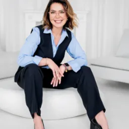 Uśmiechnięta kobieta w okularach, błękitnej koszuli i czarnej kamizelce siedzi na białym pufie, dłonie splecione, w tle białe meble.