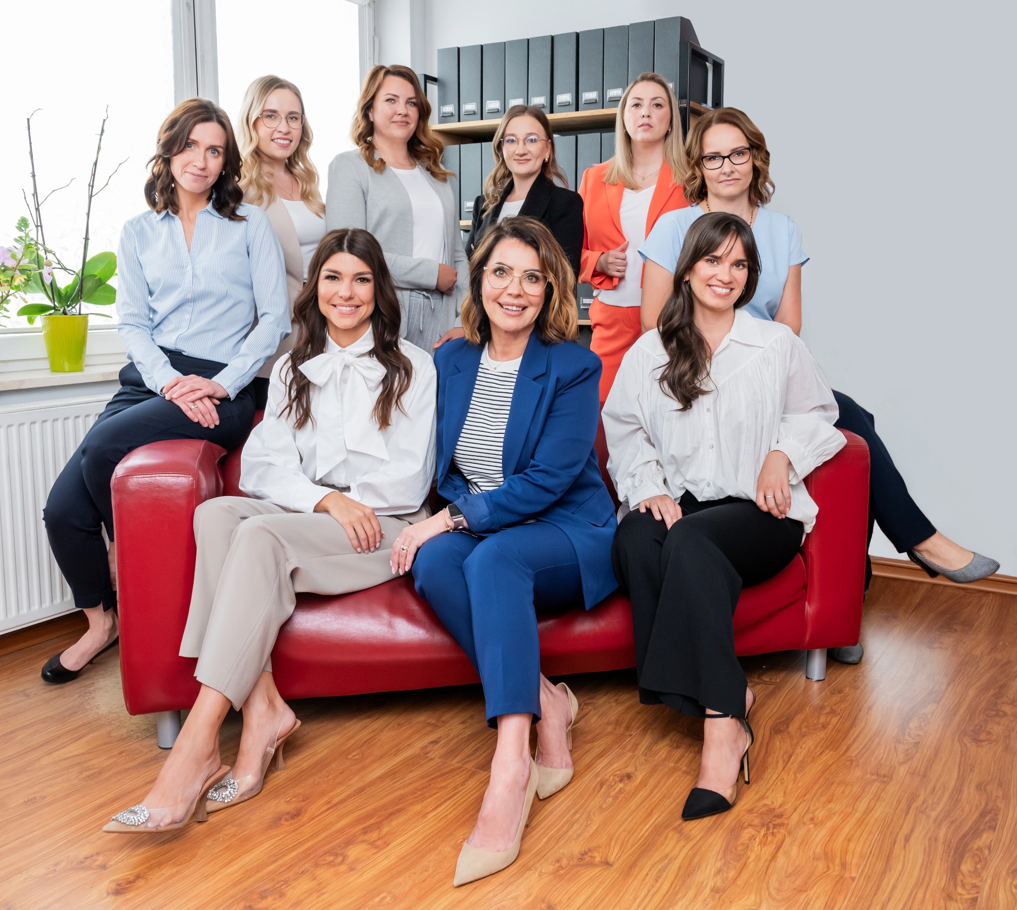 Grupowe portretowe zdjęcie dziewięciu uśmiechniętych kobiet w eleganckich strojach, siedzących na czerwonej sofie w biurze z regałem pełnym segregatorów w tle.