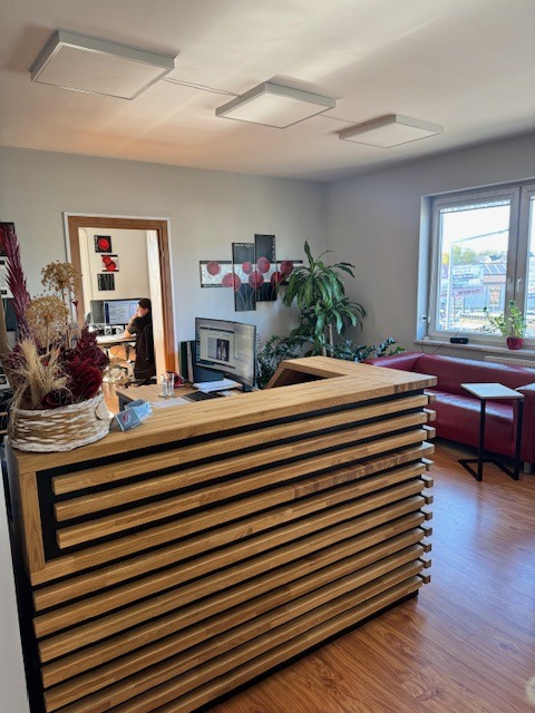 Nowoczesna recepcja z drewnianym panelem, w tle biuro z pracownikiem przy komputerze, czerwona sofa i roślina doniczkowa przy oknie.