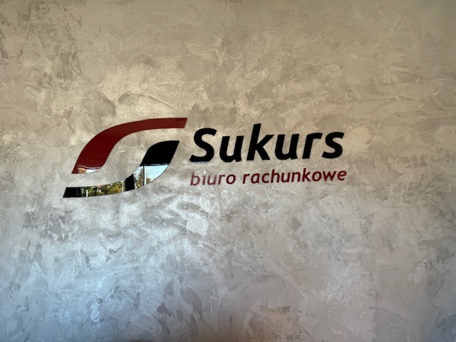 Logo firmy 'Sukurs biuro rachunkowe' umieszczone na ścianie o fakturze przypominającej beton architektoniczny, z elementami w kolorze czerwonym i czarnym oraz odbiciem otoczenia w niektórych...