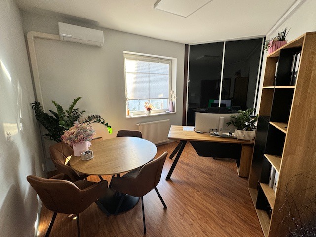 Jasne wnętrze biura z okrągłym stołem, krzesłami z brązowej skóry, biurkiem z komputerem i regałem na dokumenty, oświetlone naturalnym światłem z okna.