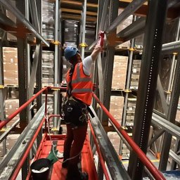 ALPI.ONE USŁUGI ALPINISTYCZNE IGOR JÄKEL - Pracownik w kasku i kamizelce odblaskowej, zabezpieczony uprzężą, na podnośniku maluje metalową konstrukcję regałów magazynowych wewnątrz hali.