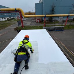 ALPI.ONE USŁUGI ALPINISTYCZNE IGOR JÄKEL - Pracownik w uprzęży i kasku maluje na biało płaski dach przemysłowy, widoczne żółte rury i budynek w tle.