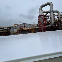 ALPI.ONE USŁUGI ALPINISTYCZNE IGOR JÄKEL - Biała, błyszcząca powierzchnia dachu przemysłowego, z widocznymi śladami po malowaniu, na tle instalacji przemysłowej z rurami i konstrukcjami stalowymi, pochmurne niebo w tle.