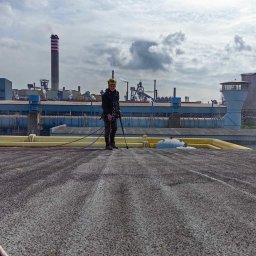 ALPI.ONE USŁUGI ALPINISTYCZNE IGOR JÄKEL - Pracownik w uprzęży na dachu przemysłowym, w tle kominy i instalacje fabryczne, widoczne ślady czyszczenia dachu.