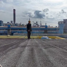 Pracownik w uprzęży na dachu przemysłowym, w tle kominy i instalacje fabryczne, widoczne ślady czyszczenia dachu.