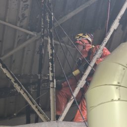 ALPI.ONE USŁUGI ALPINISTYCZNE IGOR JÄKEL - Pracownik w pomarańczowym kombinezonie i kasku, zabezpieczony linami, wykonuje prace na wysokości na konstrukcji stalowej pod dachem.