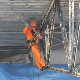 ALPI.ONE USŁUGI ALPINISTYCZNE IGOR JÄKEL - Pracownik w pomarańczowym kombinezonie, zabezpieczony linami, czyści pod wysokim ciśnieniem metalową konstrukcję dachu w hali przemysłowej. Część dachu jest już oczyszczona, w tle rozłożona...