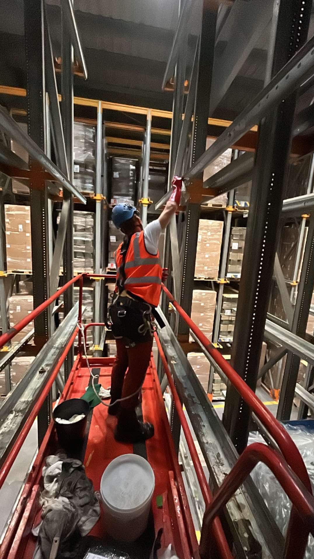 Pracownik w kasku i kamizelce odblaskowej, zabezpieczony uprzężą, na podnośniku maluje metalową konstrukcję regałów magazynowych wewnątrz hali.