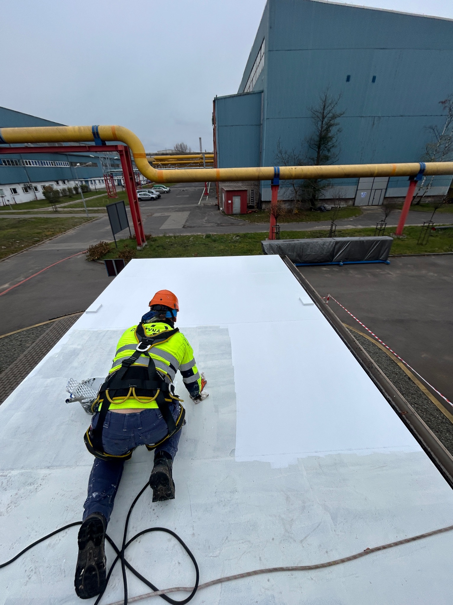 Pracownik w uprzęży i kasku maluje na biało płaski dach przemysłowy, widoczne żółte rury i budynek w tle.