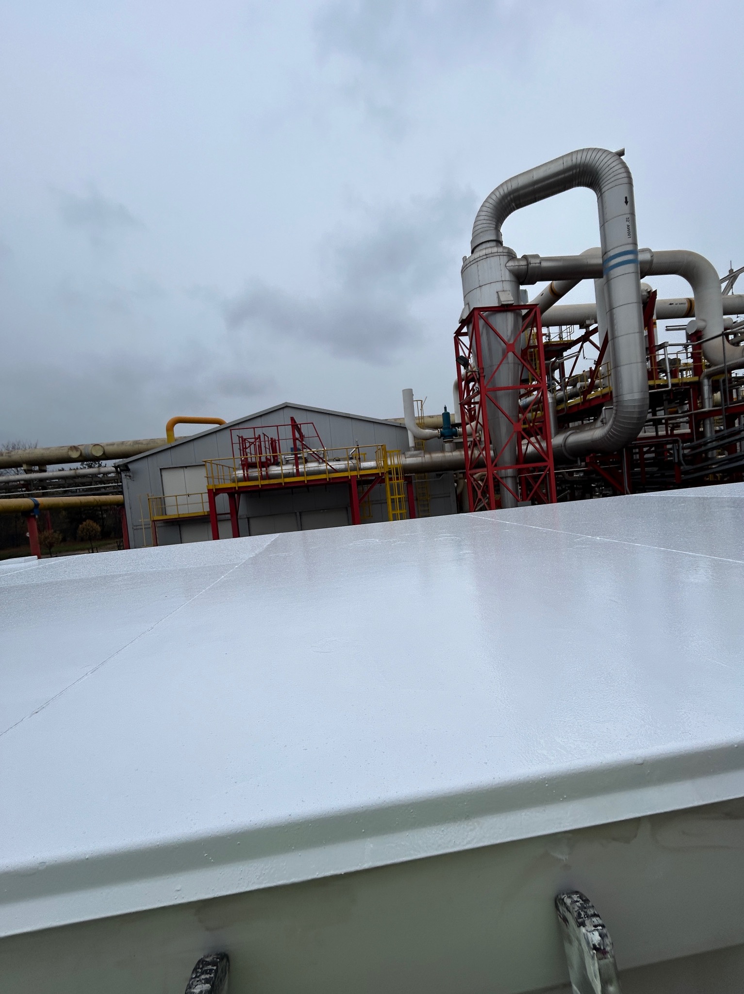 Biała, błyszcząca powierzchnia dachu przemysłowego, z widocznymi śladami po malowaniu, na tle instalacji przemysłowej z rurami i konstrukcjami stalowymi, pochmurne niebo w tle.