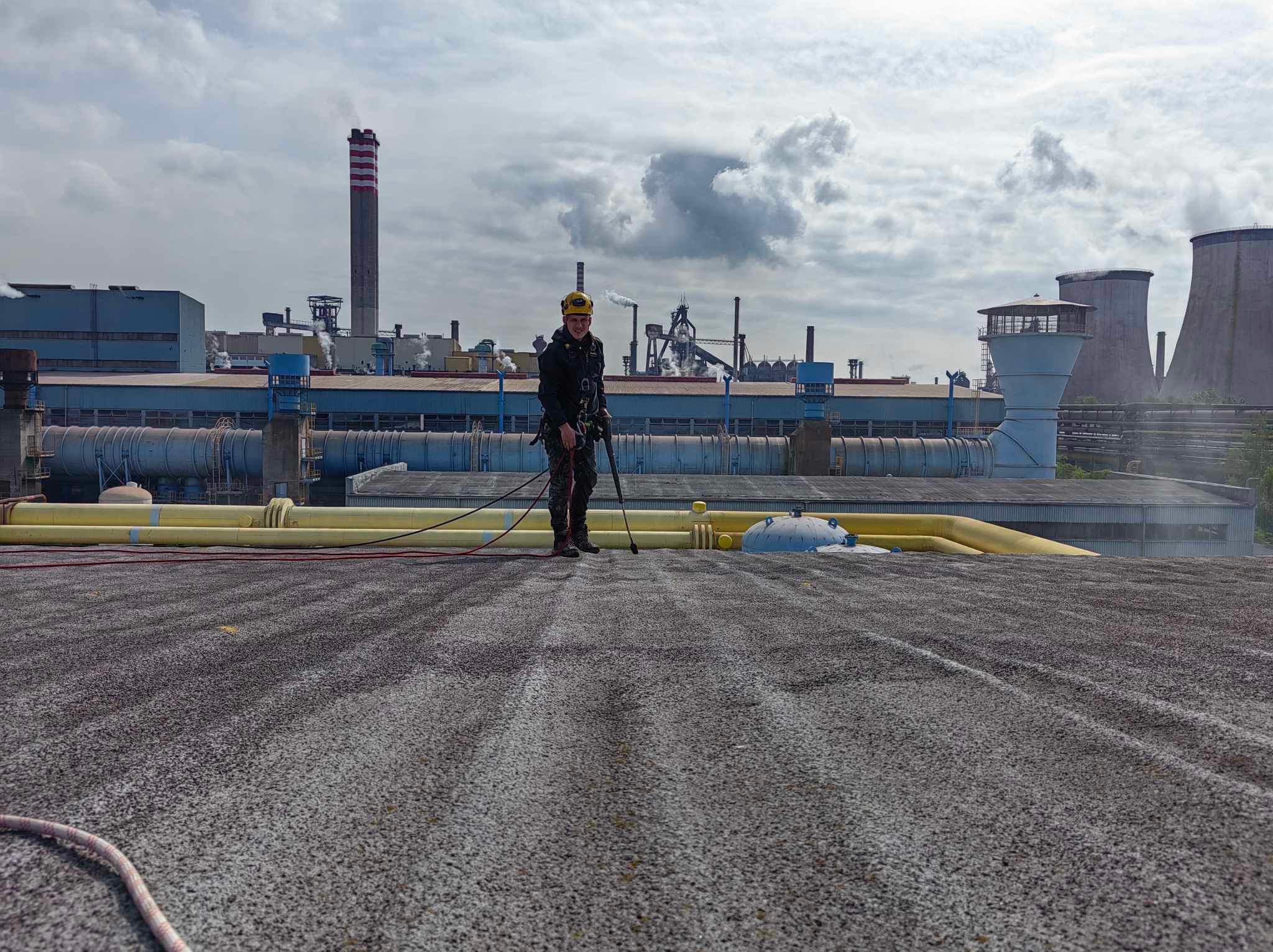 Pracownik w uprzęży na dachu przemysłowym, w tle kominy i instalacje fabryczne, widoczne ślady czyszczenia dachu.