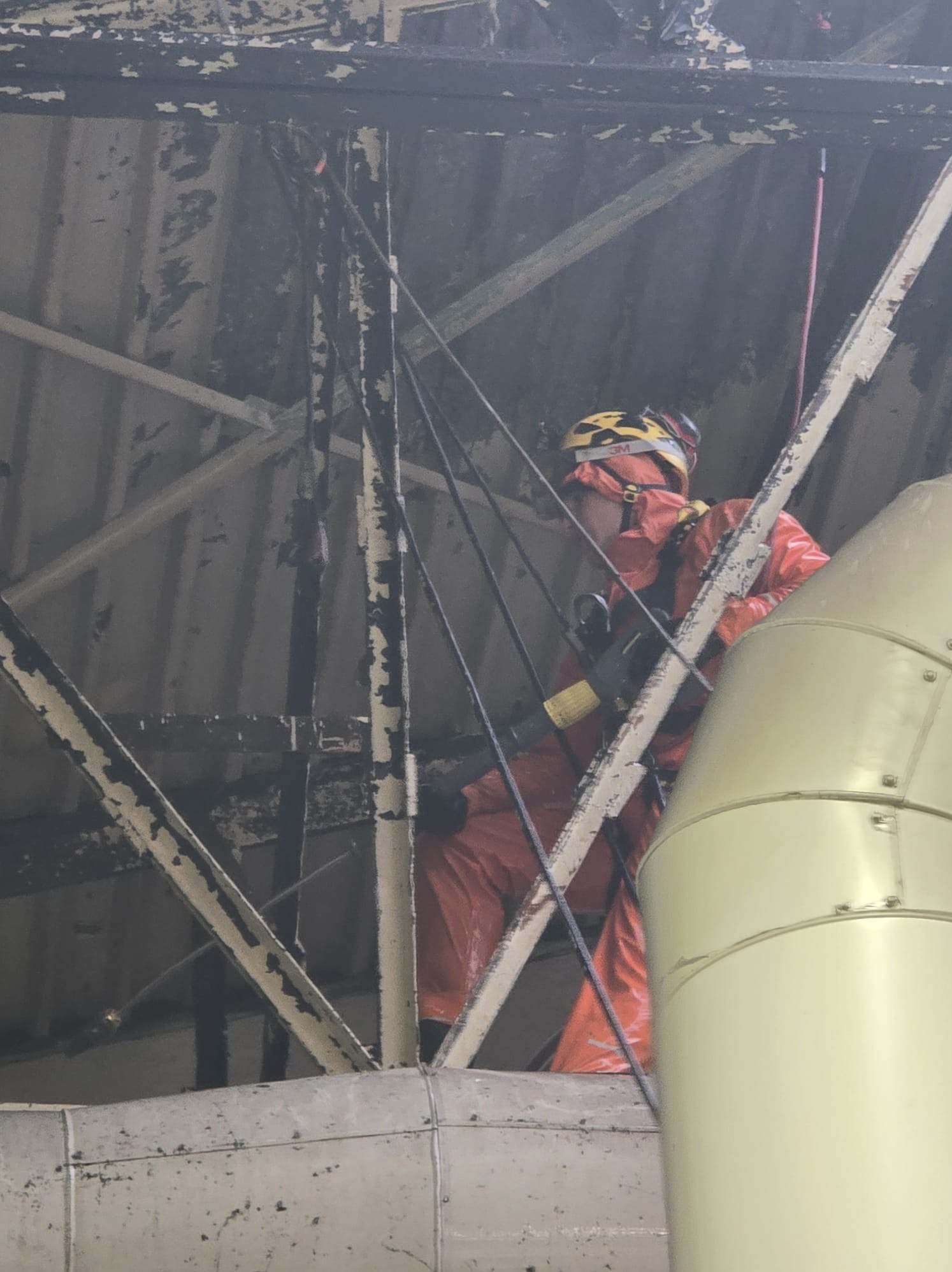 Pracownik w pomarańczowym kombinezonie i kasku, zabezpieczony linami, wykonuje prace na wysokości na konstrukcji stalowej pod dachem.