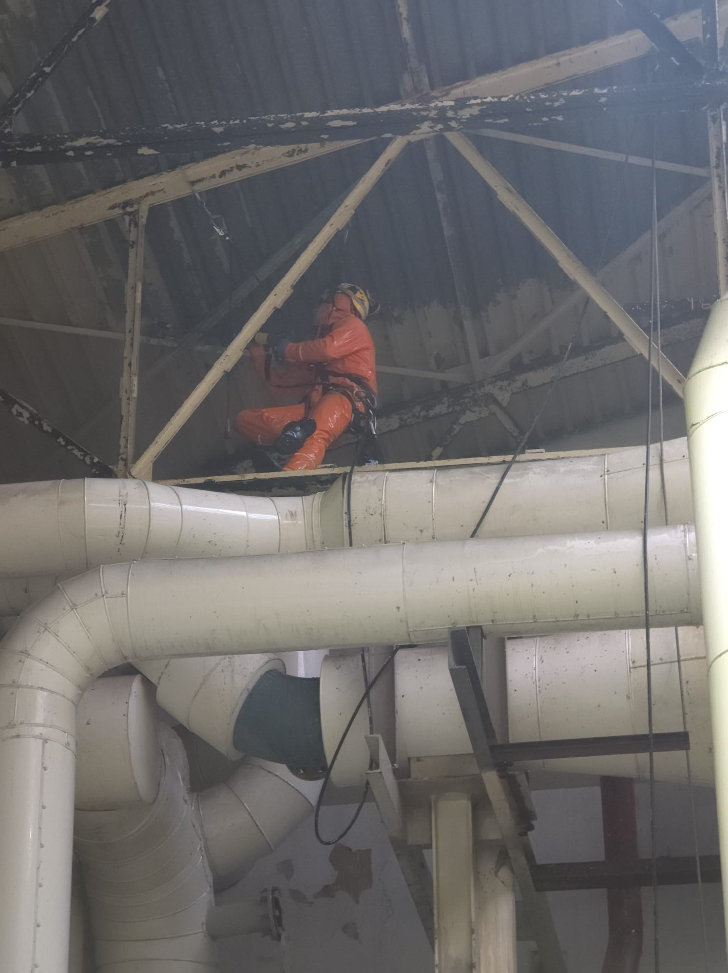 Pracownik w pomarańczowym kombinezonie i kasku, zabezpieczony linami, siedzi na konstrukcji pod dachem, prawdopodobnie podczas inspekcji lub drobnych napraw. Widoczne rury wentylacyjne i stalowa...