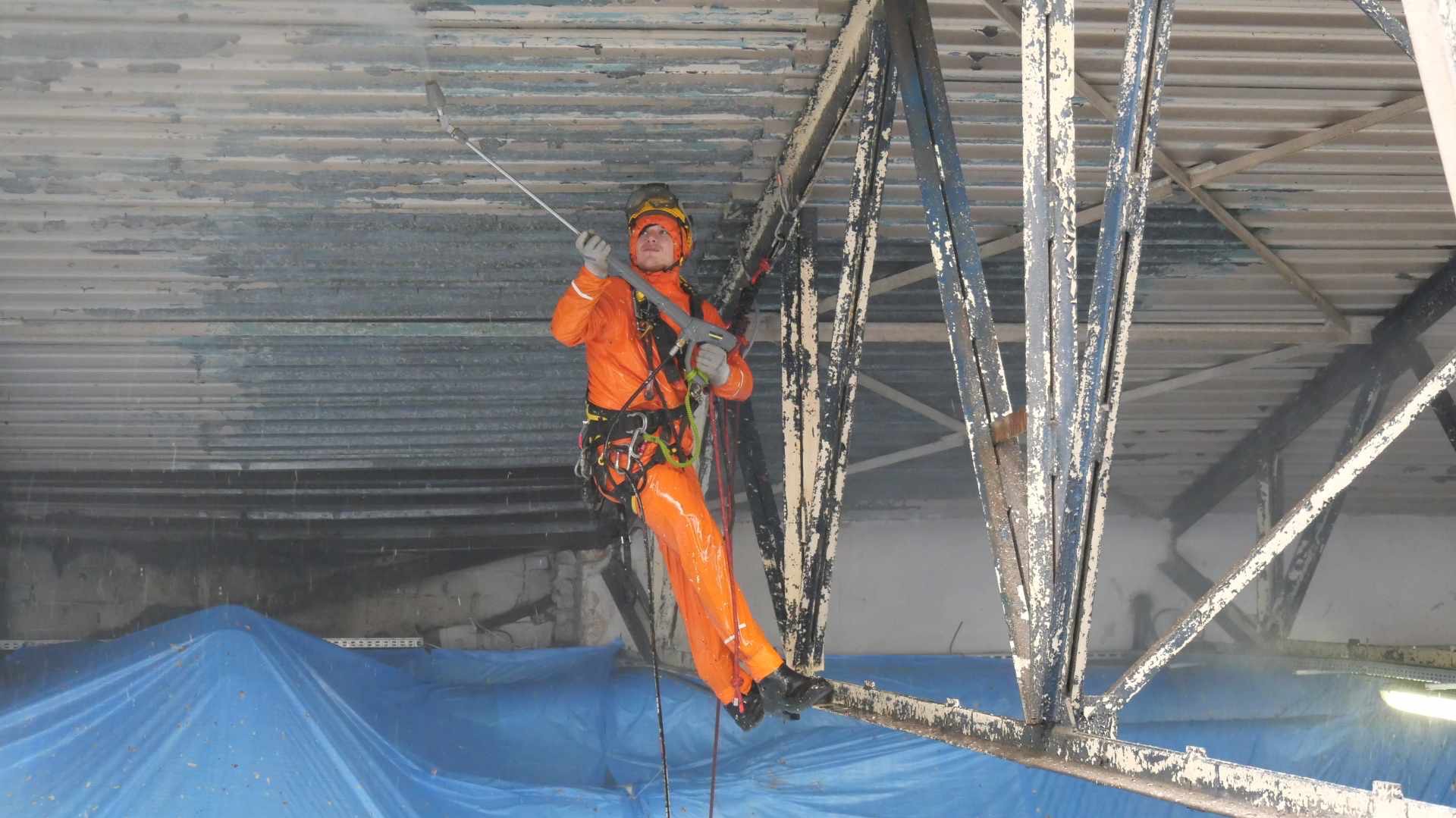 Pracownik w pomarańczowym kombinezonie, zabezpieczony linami, czyści pod wysokim ciśnieniem metalową konstrukcję dachu w hali przemysłowej. Część dachu jest już oczyszczona, w tle rozłożona...