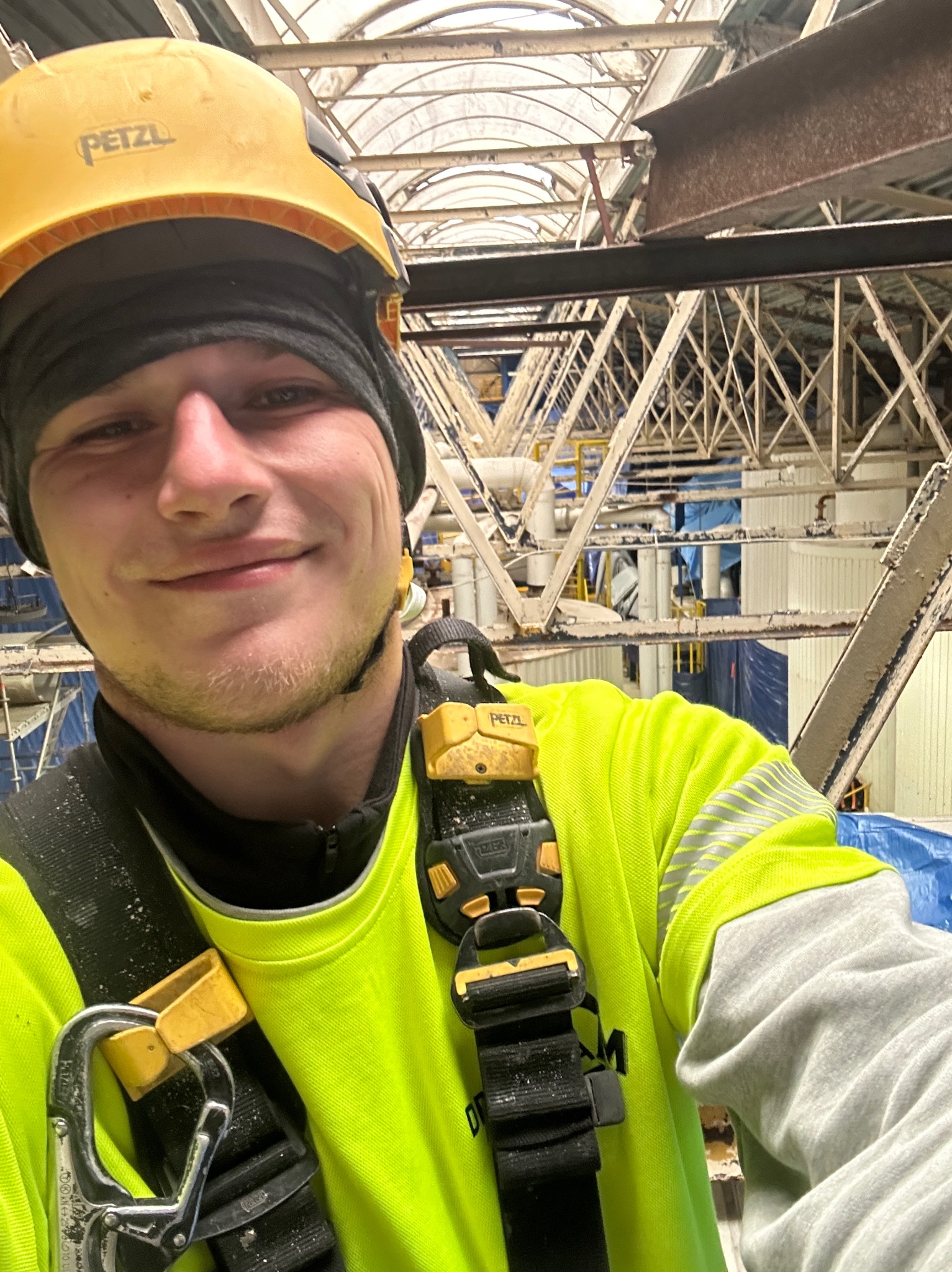 Uśmiechnięty pracownik w kasku Petzl i uprzęży, wykonujący prace na wysokości na konstrukcji stalowej z elementami dachu.
