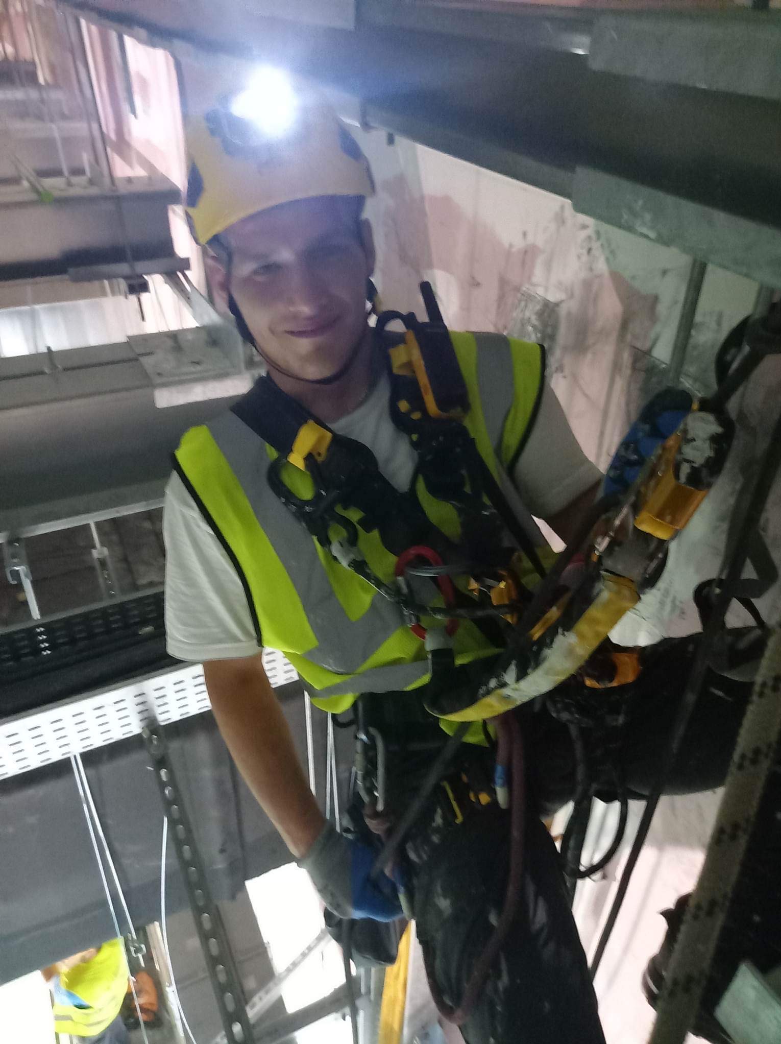 Mężczyzna w kasku z latarką i kamizelce odblaskowej, zabezpieczony linami, pracuje na wysokości wewnątrz budynku, widoczne elementy konstrukcyjne i instalacje.