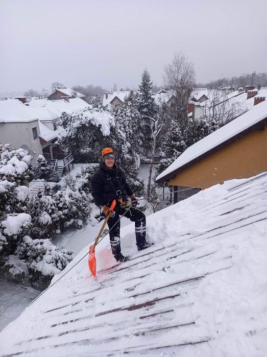 Pracownik w kasku i uprzęży odśnieża dach drewnianą łopatą na tle zasypanych śniegiem domów i drzew.
