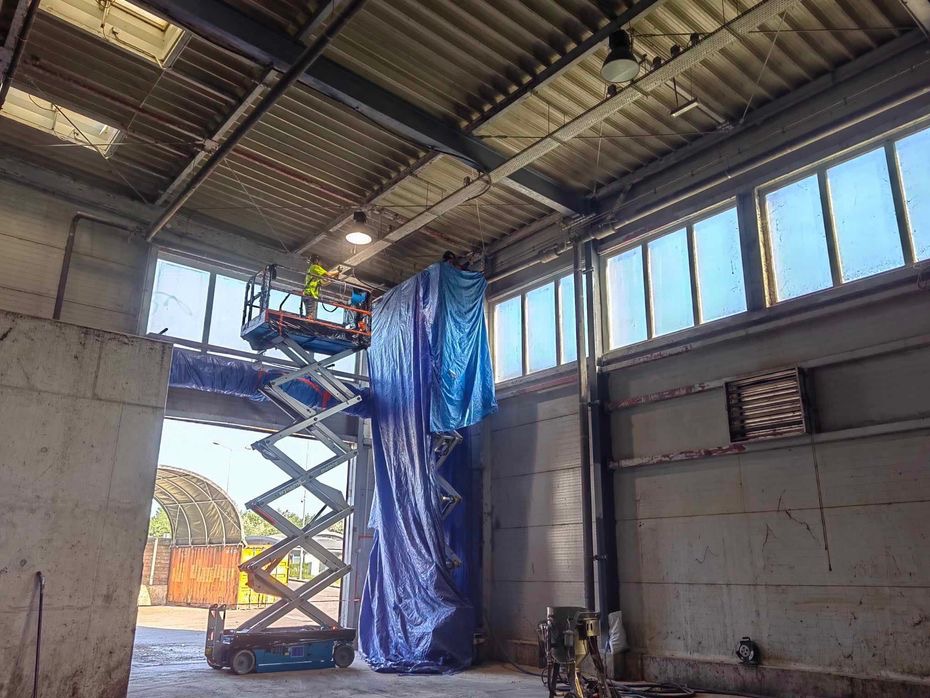 Pracownik na podnośniku nożycowym, zabezpieczony niebieską plandeką, wykonuje prace na wysokości wewnątrz hali przemysłowej, przy oknach pod sufitem.