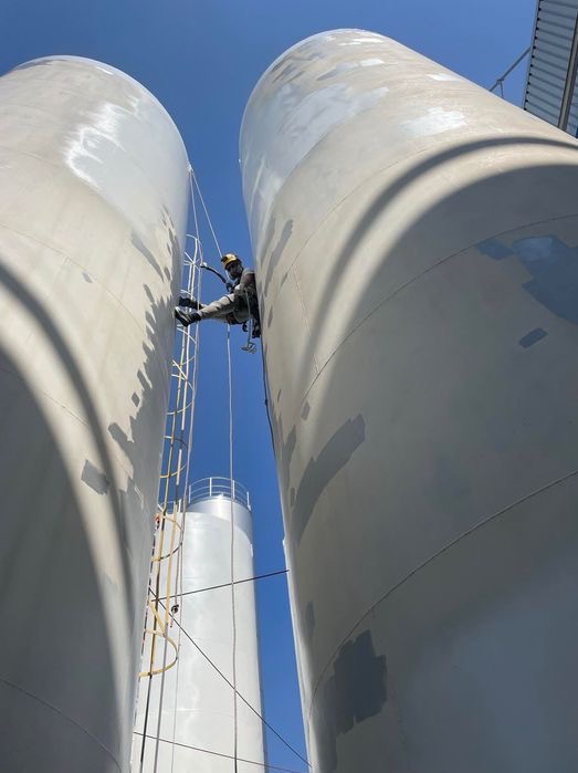 Pracownik w uprzęży, na linach, maluje silos przemysłowy na wysokości; widoczne niedomalowane fragmenty i drabina.