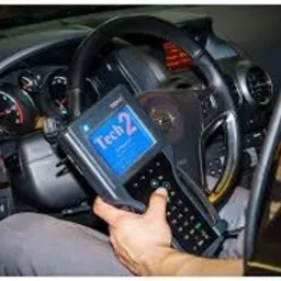 Diagnostyka komputerowa samochodu: dłoń trzyma urządzenie Tech 2 z niebieskim ekranem, widoczna kierownica i deska rozdzielcza pojazdu.