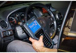 Diagnostyka komputerowa samochodu: dłoń trzyma urządzenie Tech 2 z niebieskim ekranem, widoczna kierownica i deska rozdzielcza pojazdu.