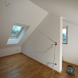 Grzegorz Bąk - Poddasze po remoncie: świeżo pomalowane białe ściany, drewniana podłoga, okno dachowe z widokiem na drzewa, widoczne gniazdka elektryczne i przedłużacz.