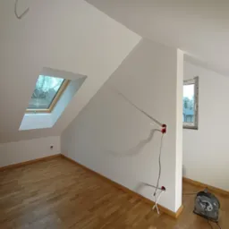 Poddasze po remoncie: świeżo pomalowane białe ściany, drewniana podłoga, okno dachowe z widokiem na drzewa, widoczne gniazdka elektryczne i przedłużacz.