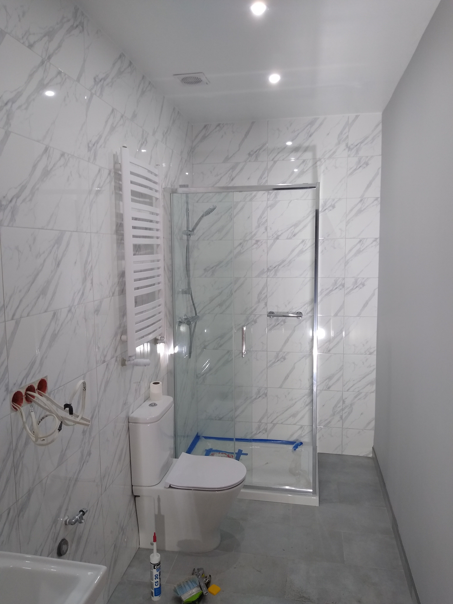 Wykończona łazienka z marmurowymi płytkami, kabiną prysznicową ze szklanymi drzwiami przesuwnymi, grzejnikiem drabinkowym, toaletą i widocznymi przewodami elektrycznymi w ścianie.