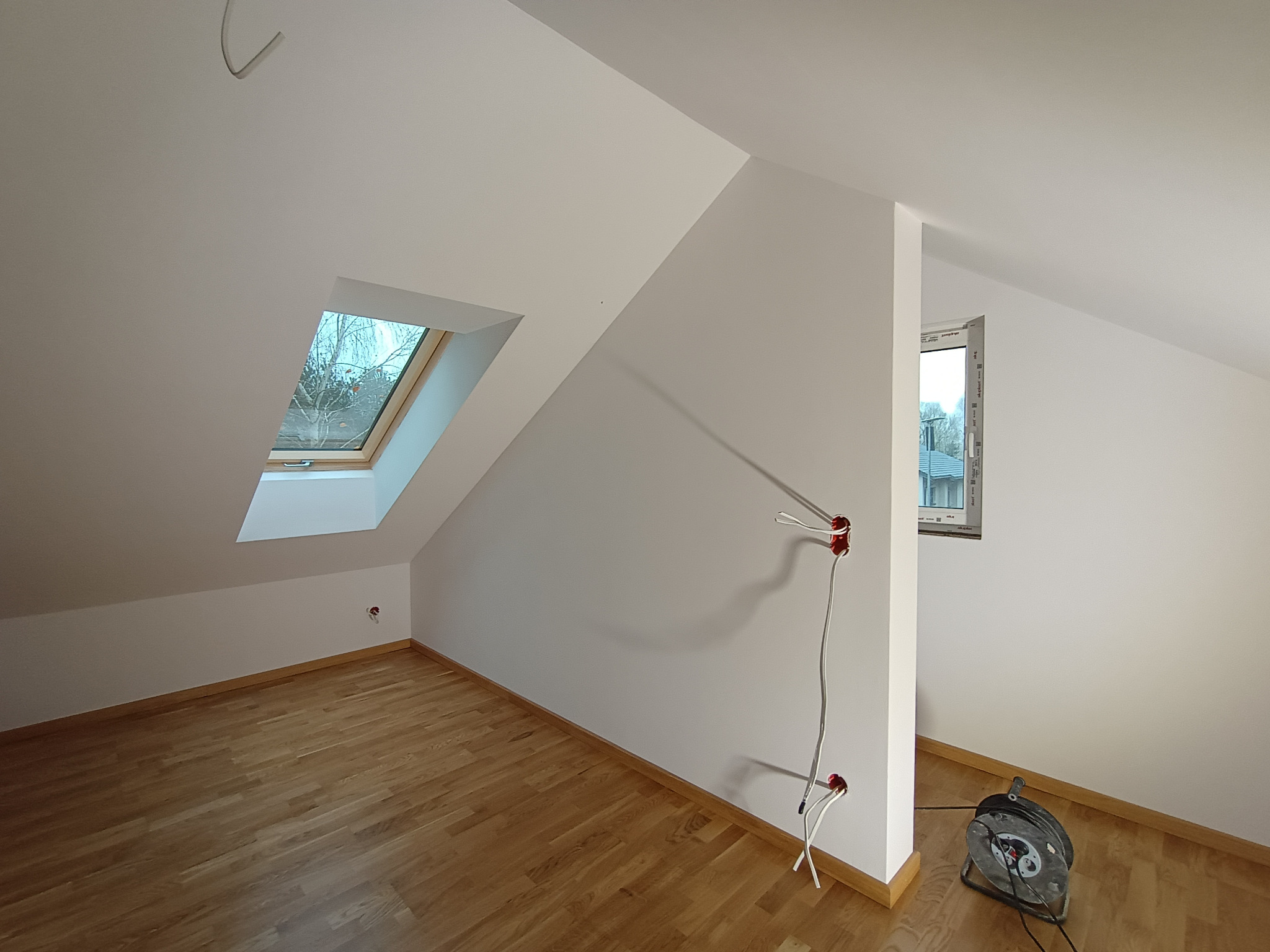 Poddasze po remoncie: świeżo pomalowane białe ściany, drewniana podłoga, okno dachowe z widokiem na drzewa, widoczne gniazdka elektryczne i przedłużacz.
