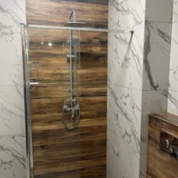 Nowoczesna kabina prysznicowa z panelami imitującymi drewno i marmurowe płytki. Chromowana armatura i szklane drzwi. Widoczny wentylator.