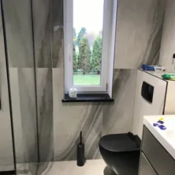 Nowoczesna łazienka z marmurowymi płytkami, czarną toaletą i umywalką z szafką. Widok na zieleń przez okno. Minimalistyczny design, eleganckie wykończenie.