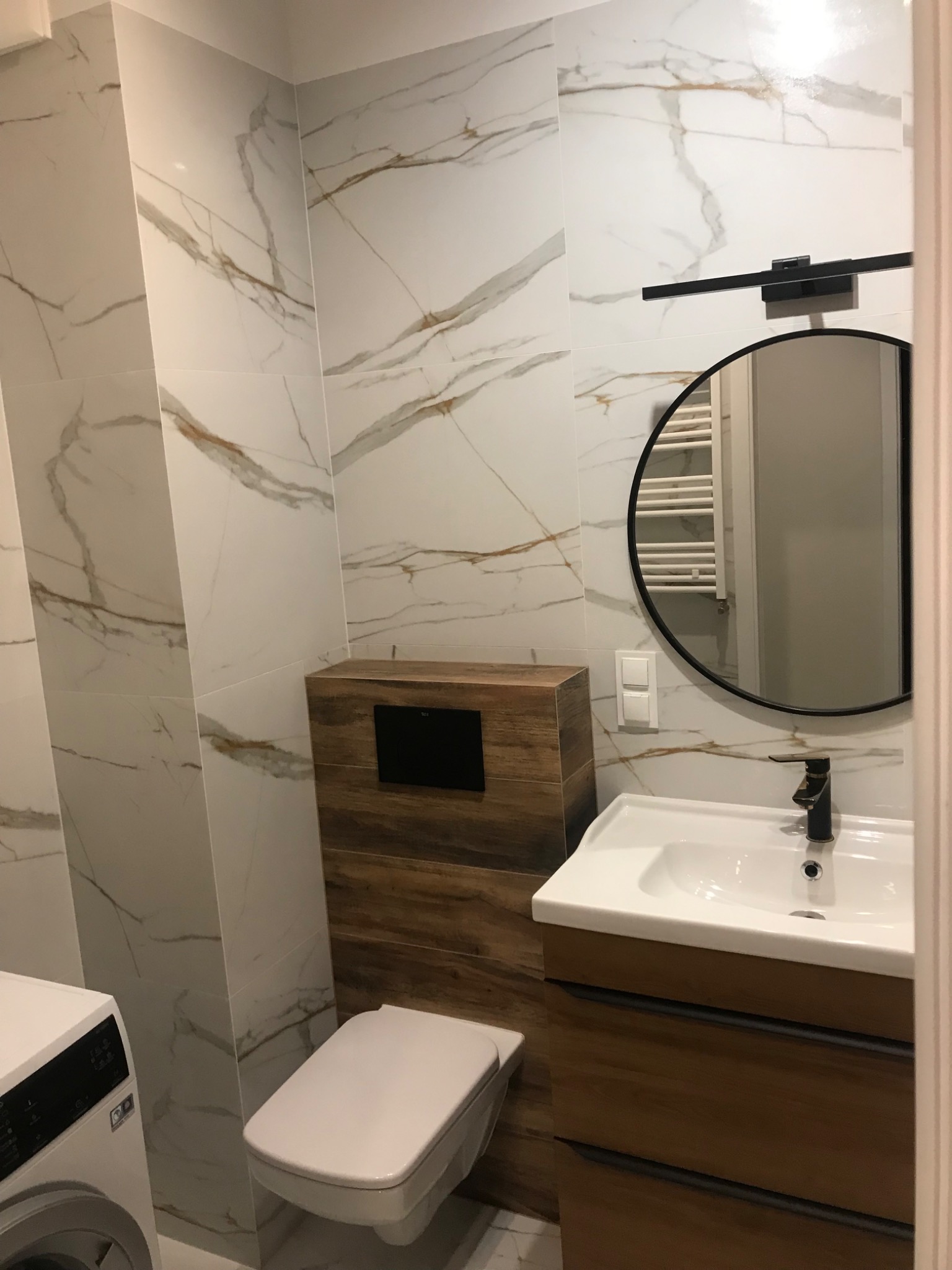 Nowoczesna łazienka z marmurowymi płytkami, drewnianą zabudową toalety i umywalki. Okrągłe lustro z czarną ramą i minimalistyczna lampa nadają elegancji.