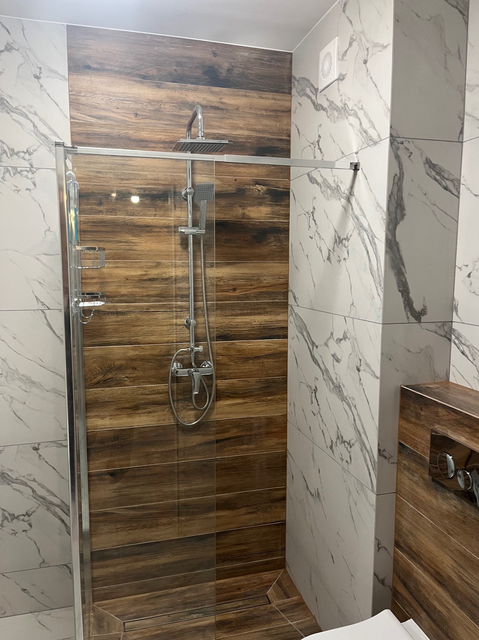 Nowoczesna kabina prysznicowa z panelami imitującymi drewno i marmurowe płytki. Chromowana armatura i szklane drzwi. Widoczny wentylator.
