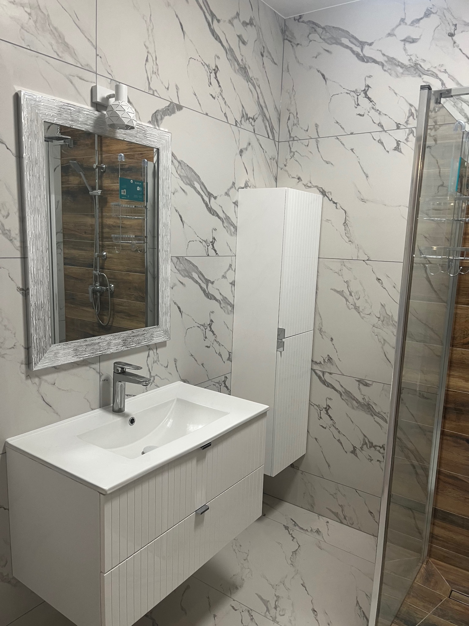 Nowoczesna łazienka z białymi płytkami imitującymi marmur, białą szafką pod umywalką i lustrem w srebrnej ramie. Widoczna kabina prysznicowa z drewnianym akcentem.